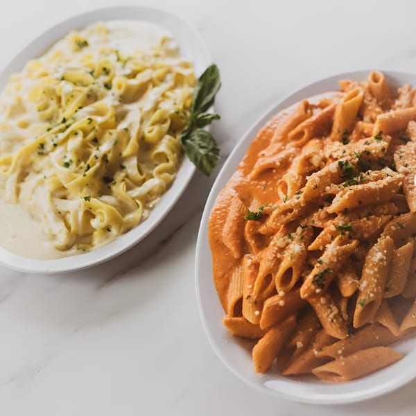 Two plates of pasta, one with creamy white sauce and the other with red sauce, on a white table.
