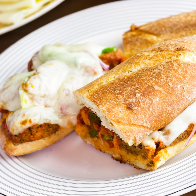 Two cheesy meatball subs on a white plate, one cut open to show the filling.