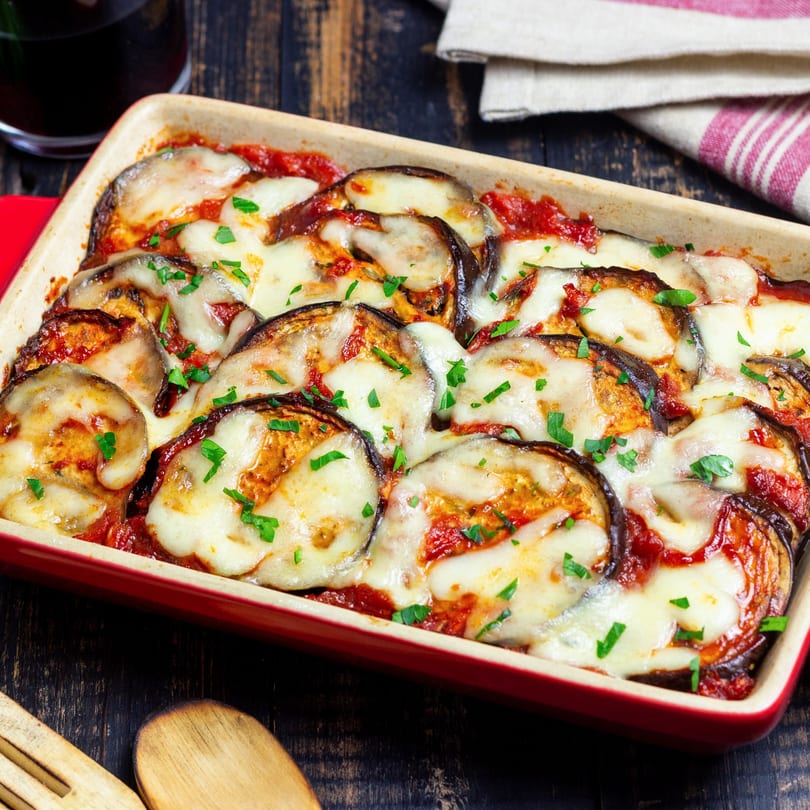 Baked eggplant parmesan with melted mozzarella cheese, tomato sauce, and parsley in a red dish.